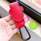 Red leather keychain with carabiner held in a hand, with colorful objects in the background.