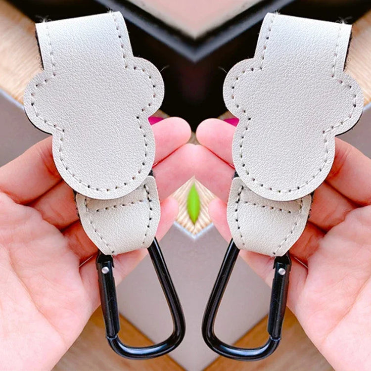 Two beige leather straps with black carabiners held by hands against a neutral background.